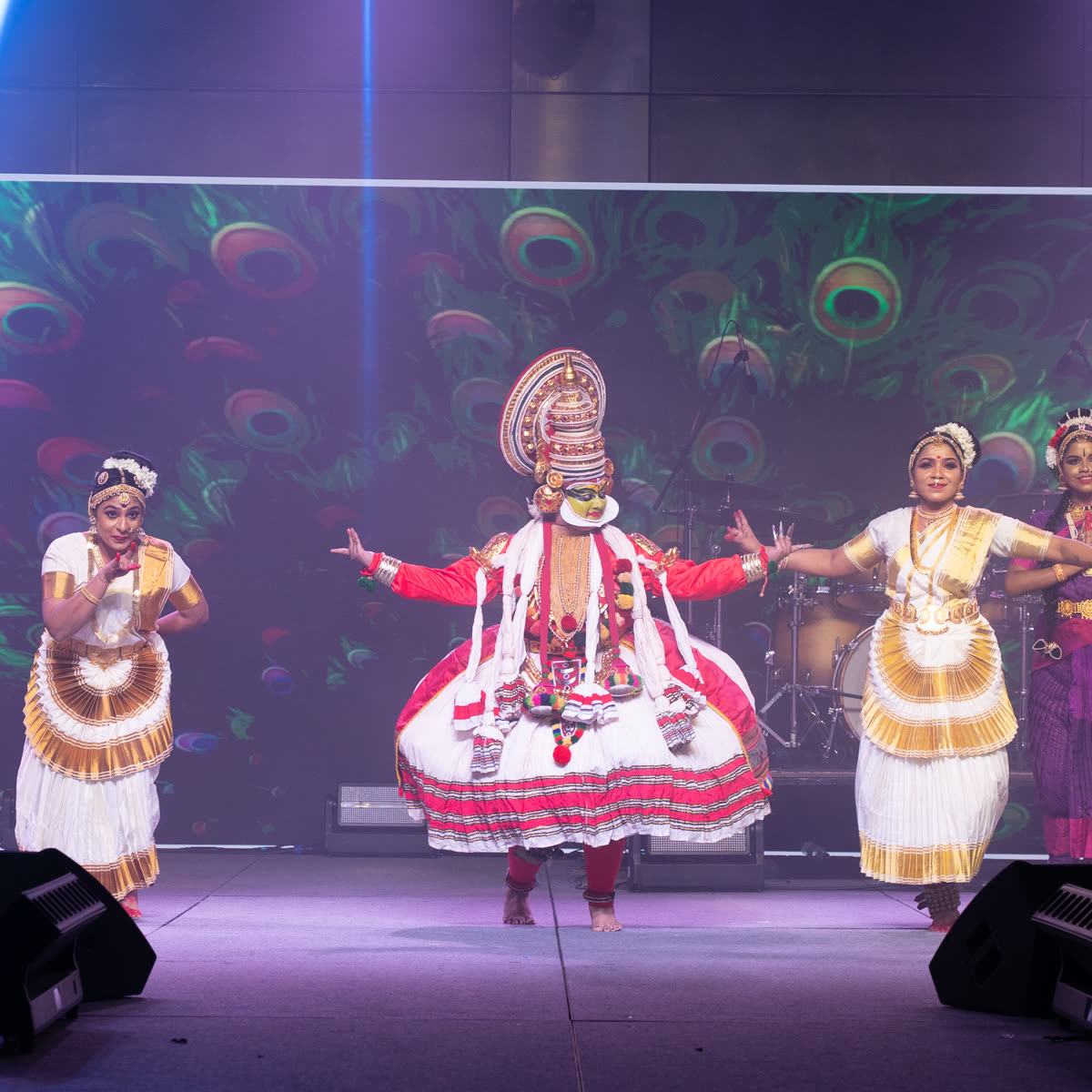 A Kathakali artist flanked by dancers against a peacock feather backdrop