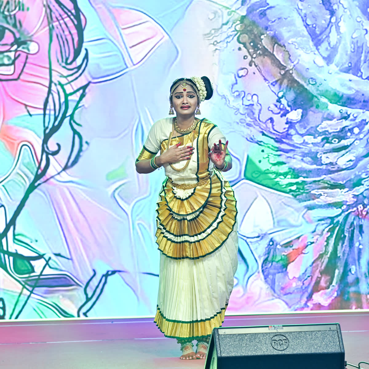 A solo Mohiniyattam dancer in white and gold mid-pose against a painted backdrop