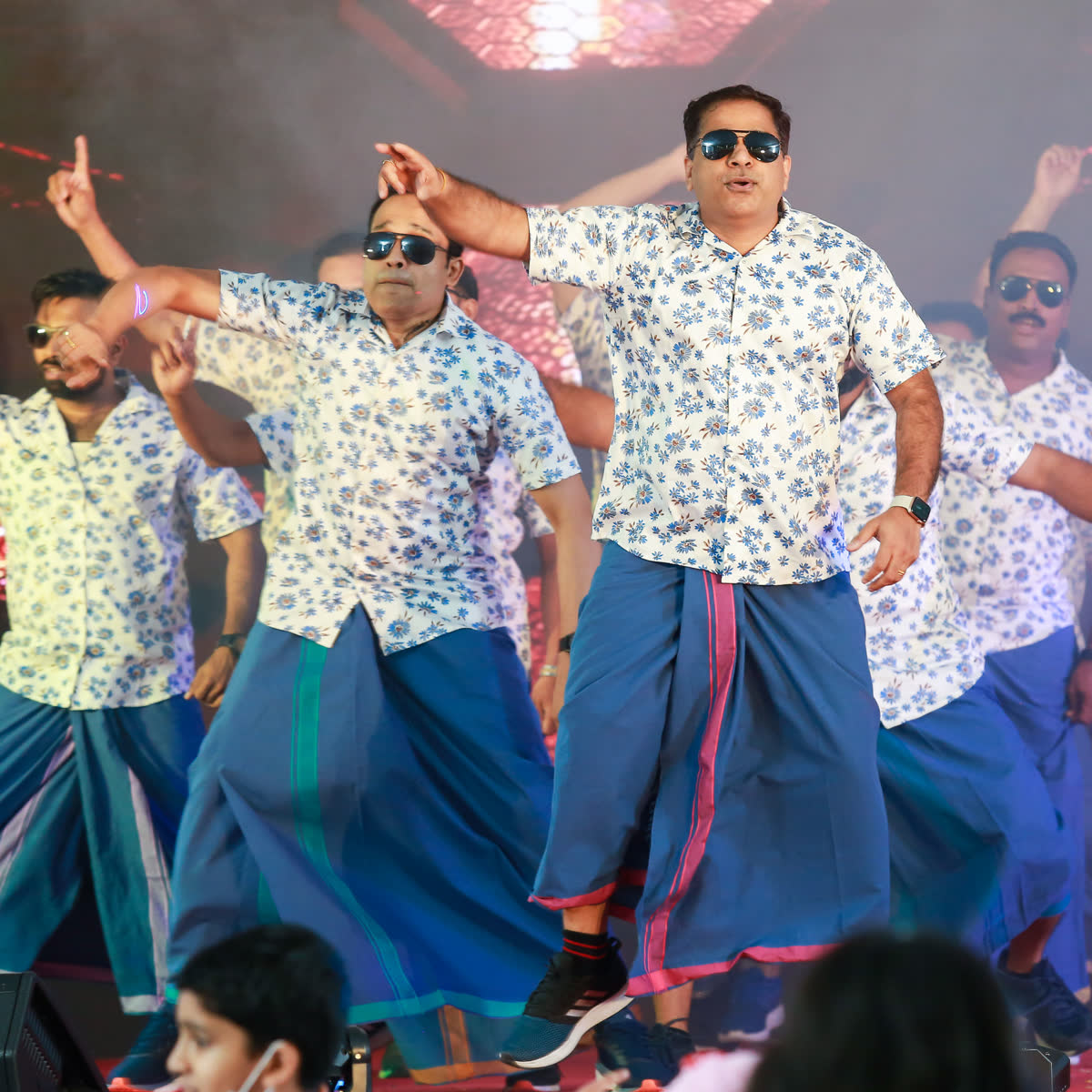 A line of men in printed shirts and blue mundus performing a group dance on stage