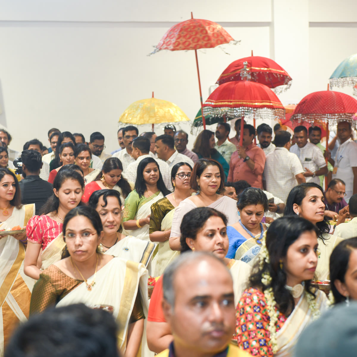 Members in cream and gold kasavu sarees walking in an Onam procession under red and white ceremonial umbrellas