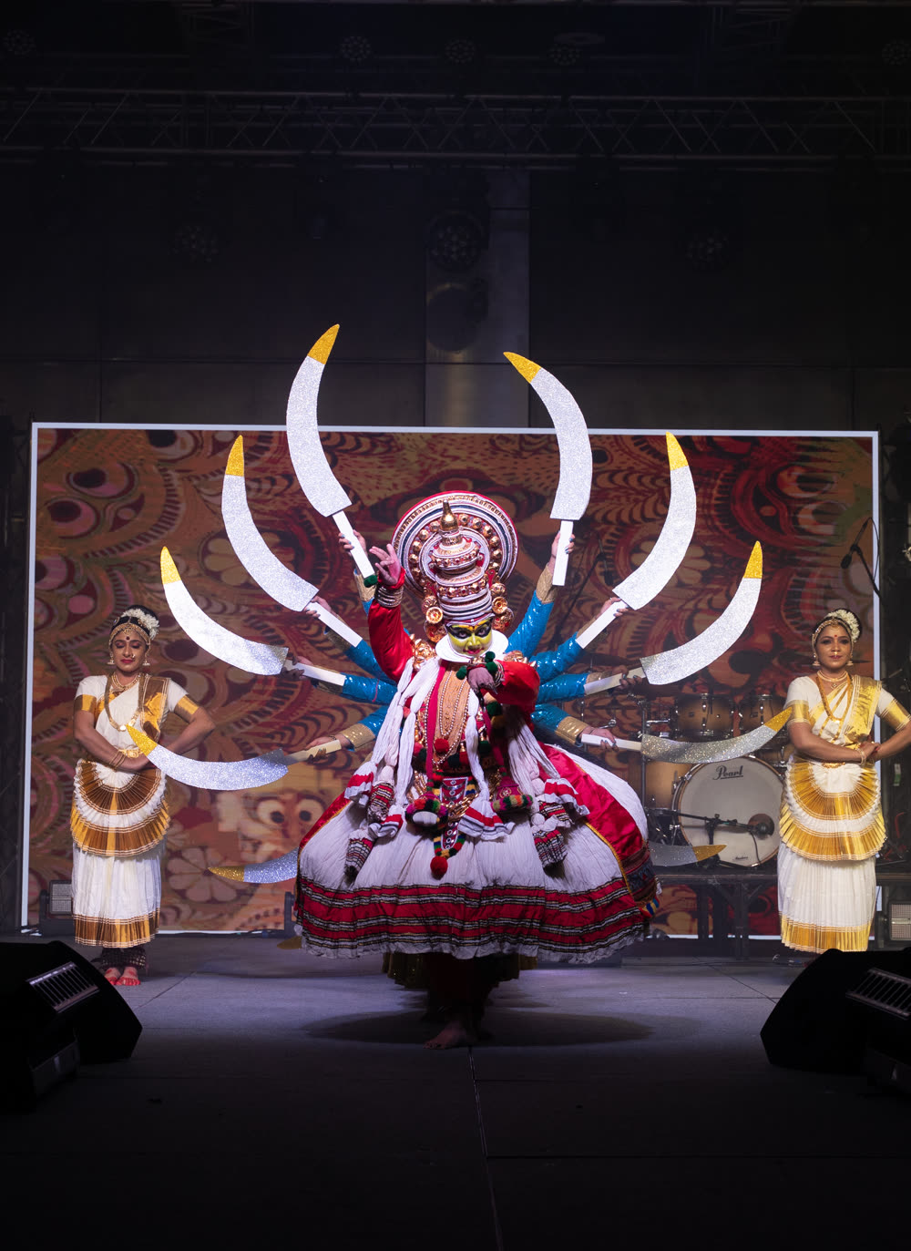 A performer in an elaborate fanned headdress and traditional costume lit against a dark stage