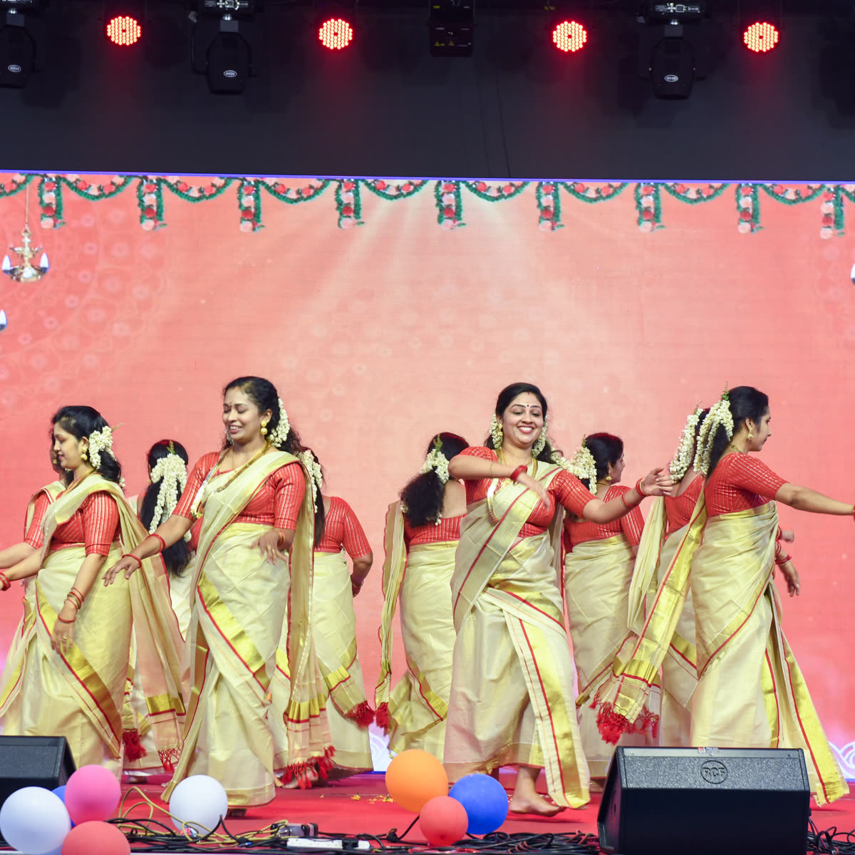 Women in kasavu sarees dancing in formation on a stage lit in pink